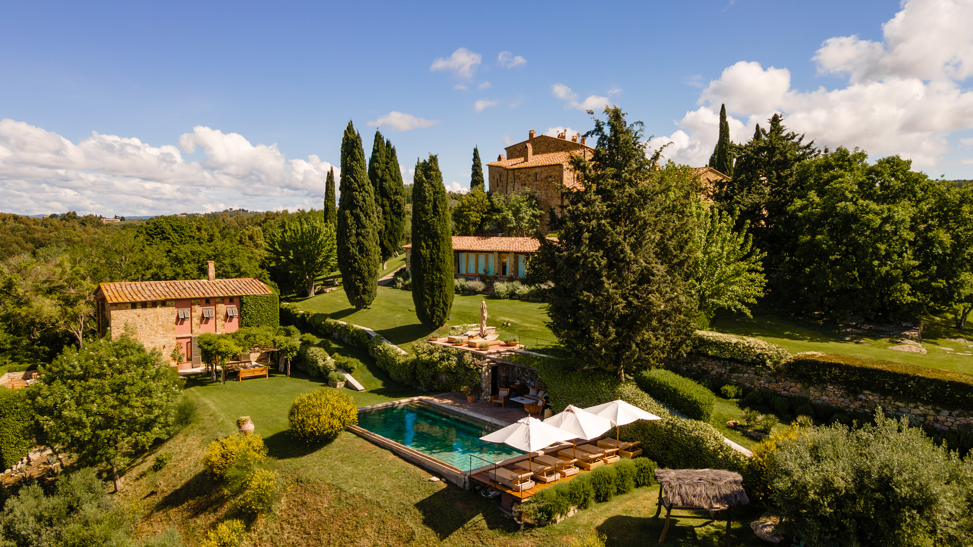 Tuscan landscape featuring Castello di Vicarello, a villa with a pool, cypress trees, and lush greenery under a blue sky.