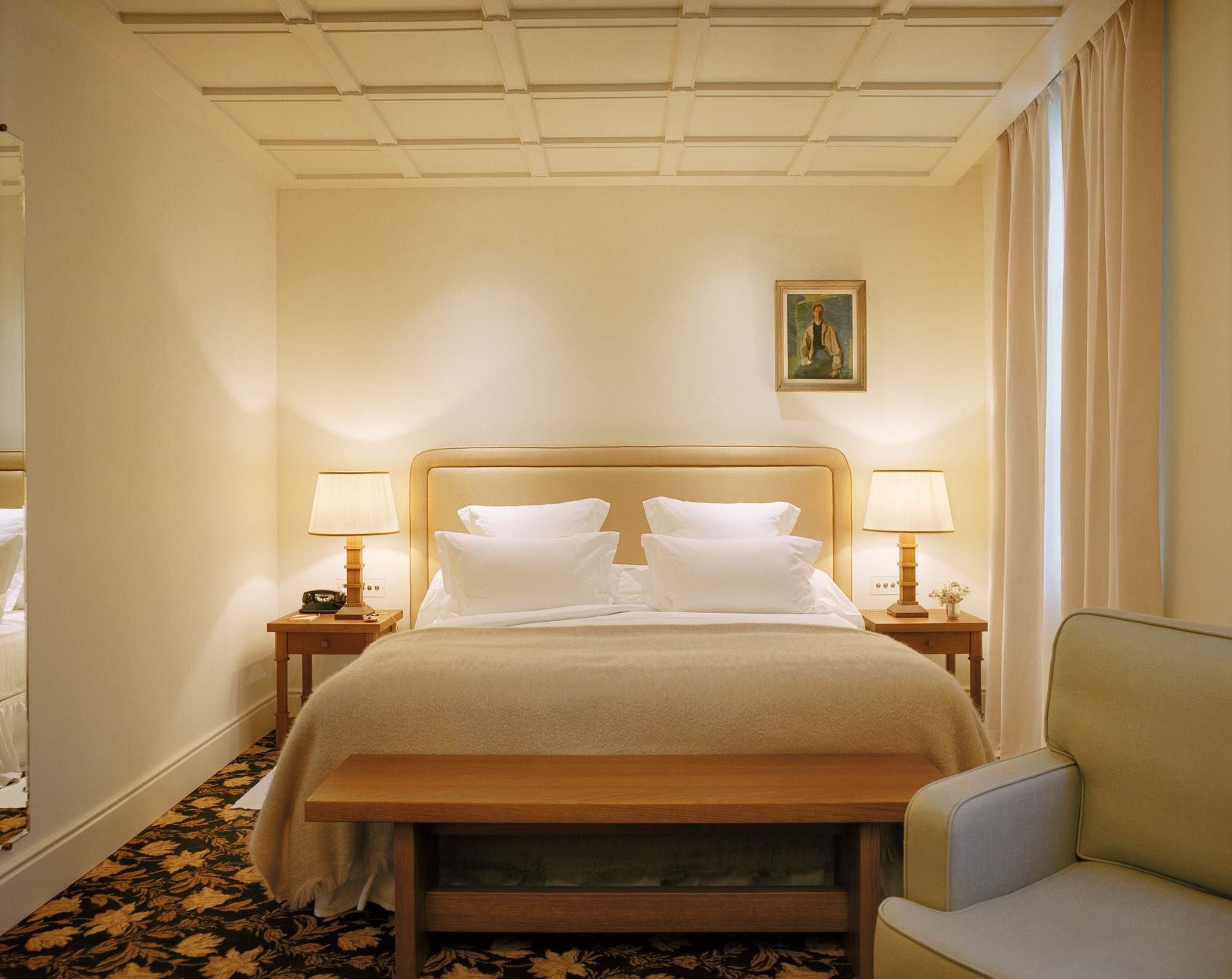 Elegant hotel room with a double bed, bedside lamps, a painting on the wall, and a cozy seating area.
