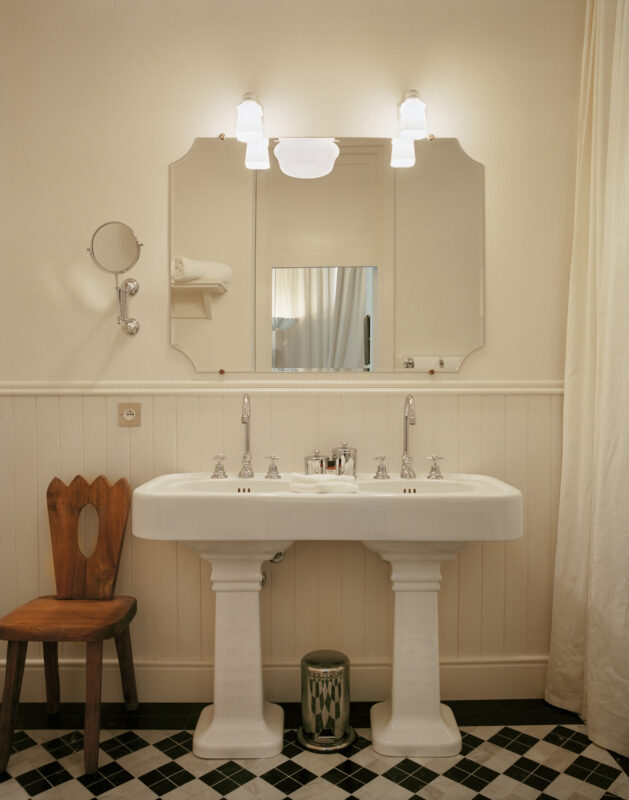 Elegant bathroom featuring a white pedestal sink, vintage-style fixtures, a large mirror, and a wooden chair against a til...