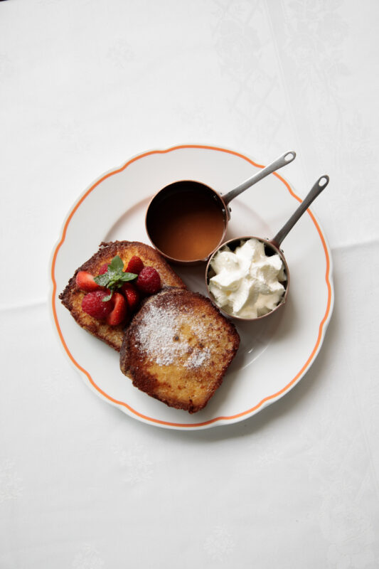 Two slices of golden-brown French toast topped with strawberries and powdered sugar, served with chocolate sauce and whipp...