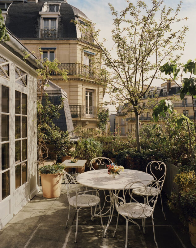 Charming terrace with a round white table and chairs, surrounded by greenery and a view of a classic building.