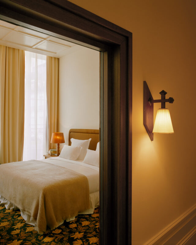 Cozy hotel room with a bed, warm lighting, and soft furnishings, viewed through a doorway.