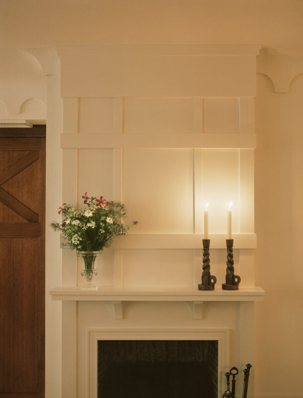 Minimalist fireplace with a white mantel, two lit candles, and a vase of flowers against a softly lit wall.