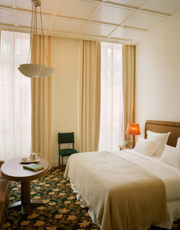 Elegant hotel room with a bed, round table, and green chair, featuring soft lighting and large windows with sheer curtains.