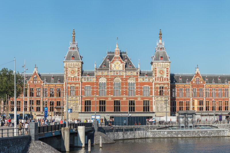 Amsterdam Centraal station's ornate facade with towers, set against a clear blue sky, viewed from the waterfront.
