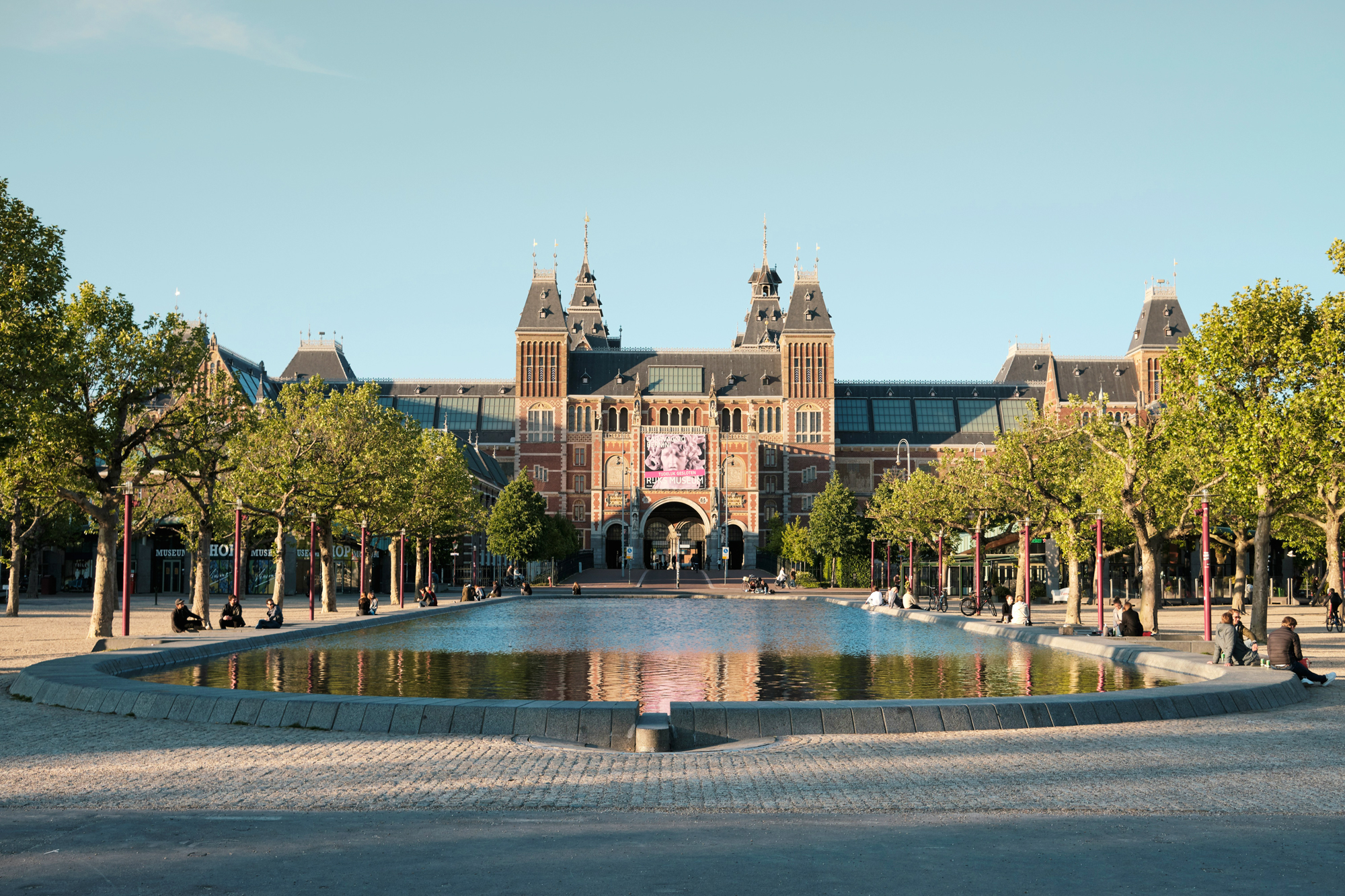 Rijksmuseum facade reflected in a pond, surrounded by trees and visitors enjoying the sunny day.