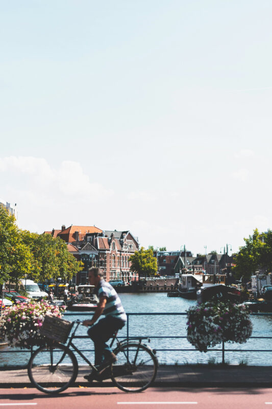A cyclist rides along a waterfront path, with colorful flowers and buildings lining the canal in the background.