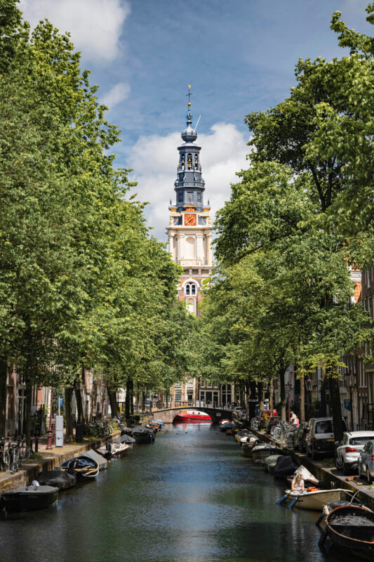 Canal lined with trees, boats, and a clock tower in the background under a partly cloudy sky.