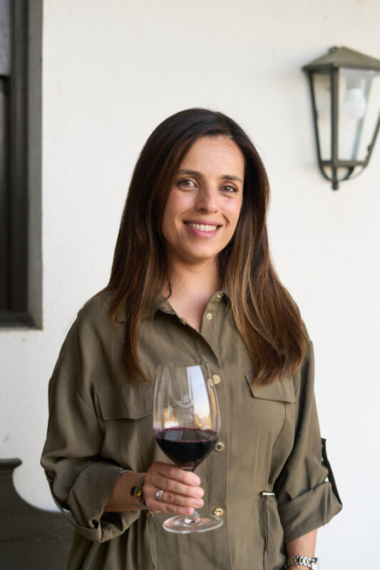 Winemaker Catia Barbeta holding a glass of red wine, smiling, in front of a white wall with a lantern.