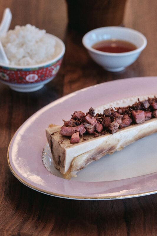 Bone marrow topped with diced meat, served on a pink plate, alongside rice and sauce in small bowls.