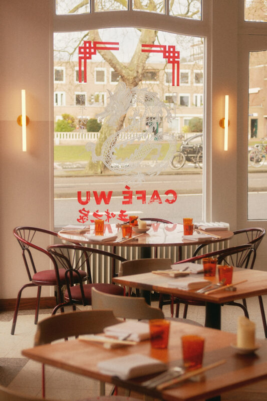 Interior of Café Wu featuring a round table with orange glasses, red chairs, and a large window displaying the café's name.