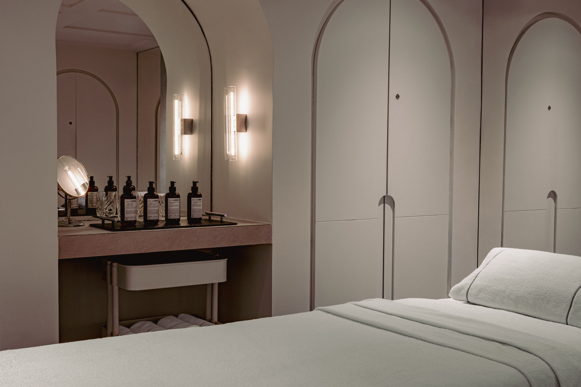 Minimalist spa interior featuring a treatment bed, illuminated mirrors, and neatly arranged skincare products on a counter.