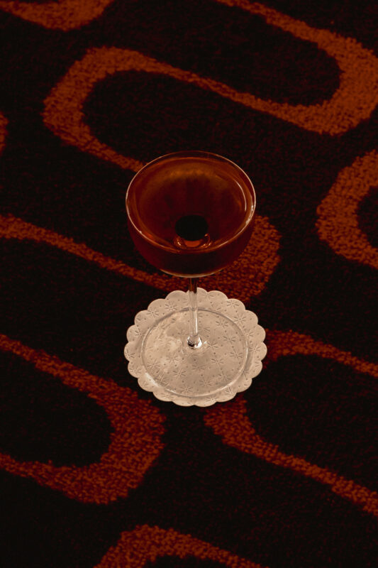 Brown cocktail glass on a decorative lace coaster, set against a patterned dark red carpet.