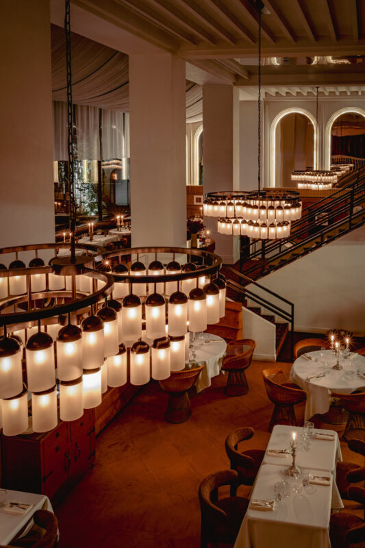 Elegant restaurant interior featuring large, modern chandeliers and neatly set tables, with a staircase in the background.