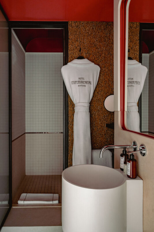 Modern bathroom featuring a white robe labeled "Hotel Experimental Marais," a round sink, and a glass shower.