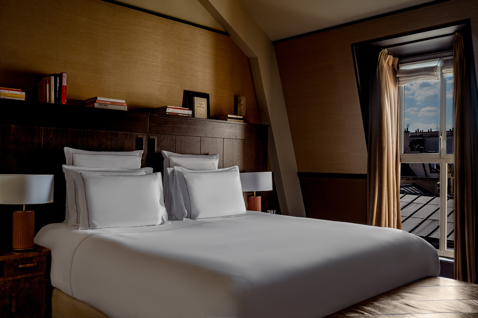 Modern bedroom with a neatly made bed, two bedside lamps, and a window showing a view of the sky and rooftops.