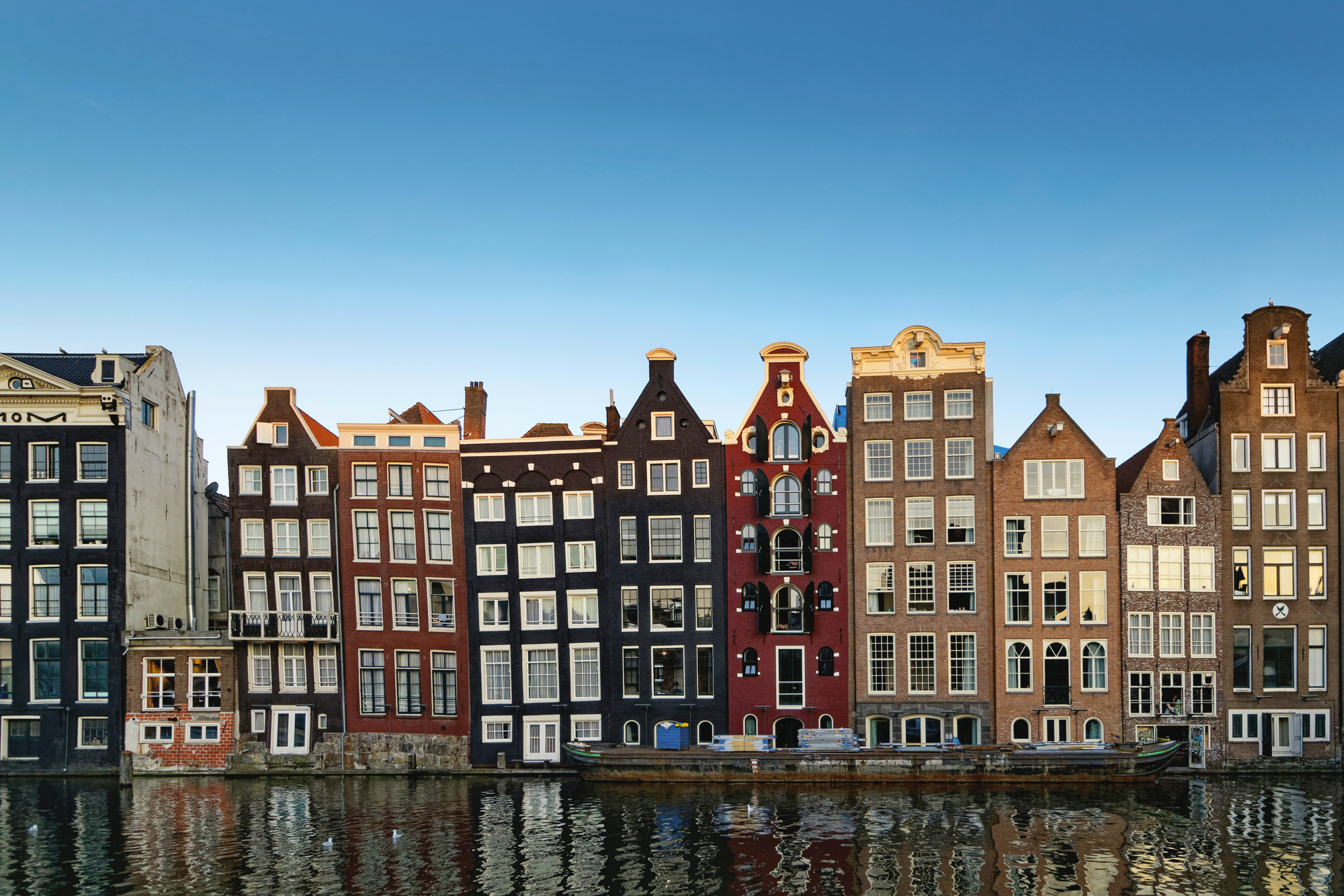 Colorful, historic buildings line a canal in Amsterdam, reflecting in the water under a clear blue sky.