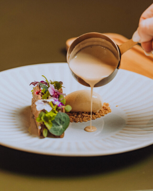 Dessert plate featuring a chocolate dessert with edible flowers, a scoop of ice cream, and sauce being poured over it.