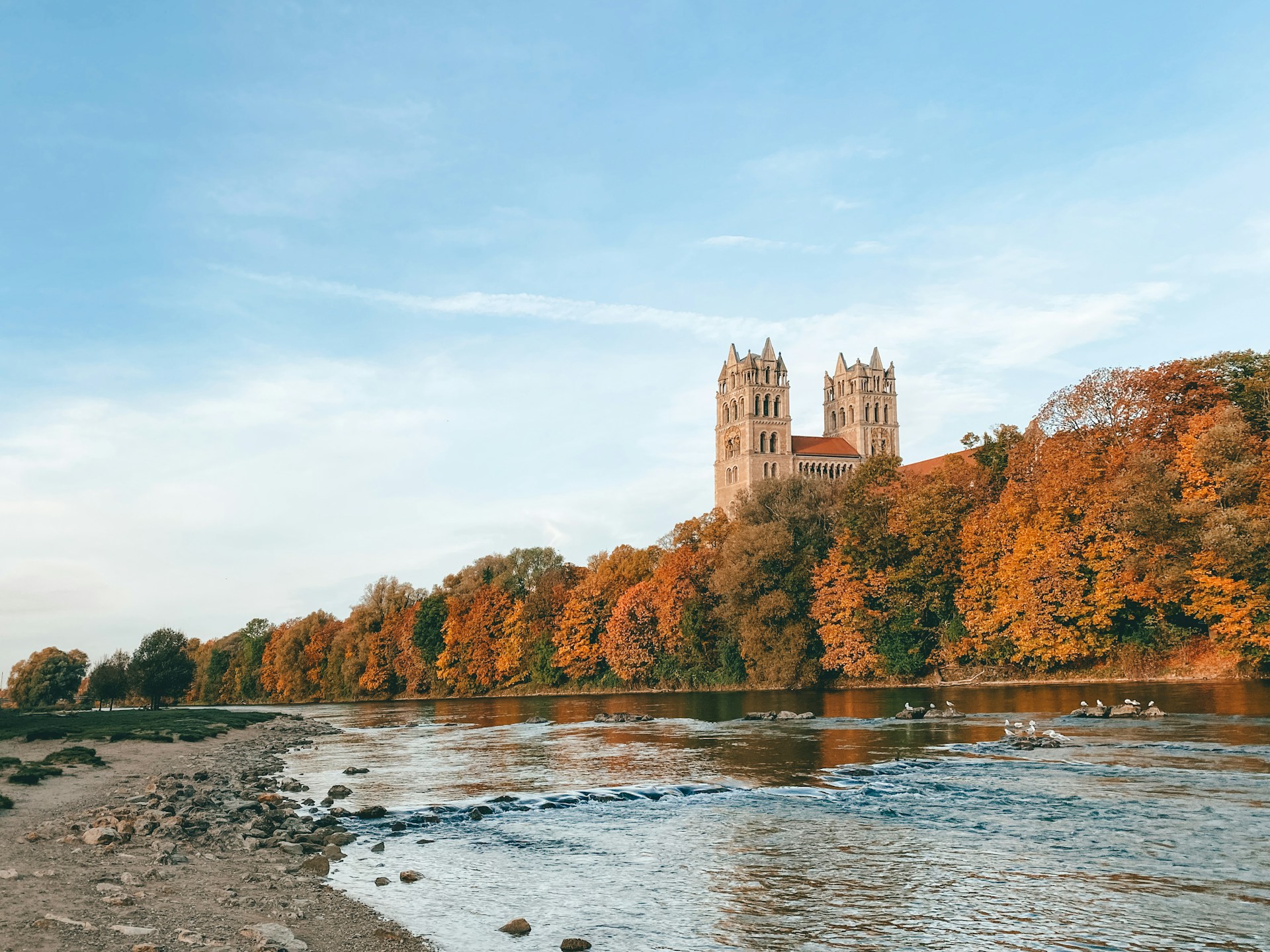 Autumn landscape featuring a river, rocky shore, and a historic building with twin towers surrounded by colorful trees.