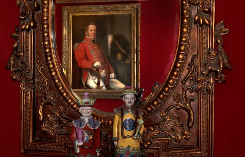 Portrait of a man in a red military uniform framed in an ornate mirror, with decorative figurines in traditional attire be...