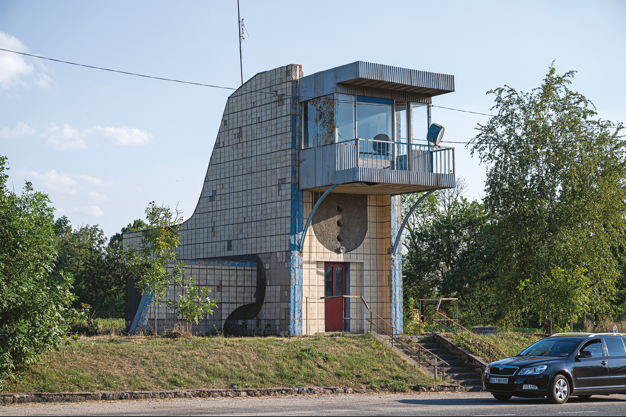 Unique architectural structure resembling a modernist building, featuring large windows and a balcony, surrounded by green...
