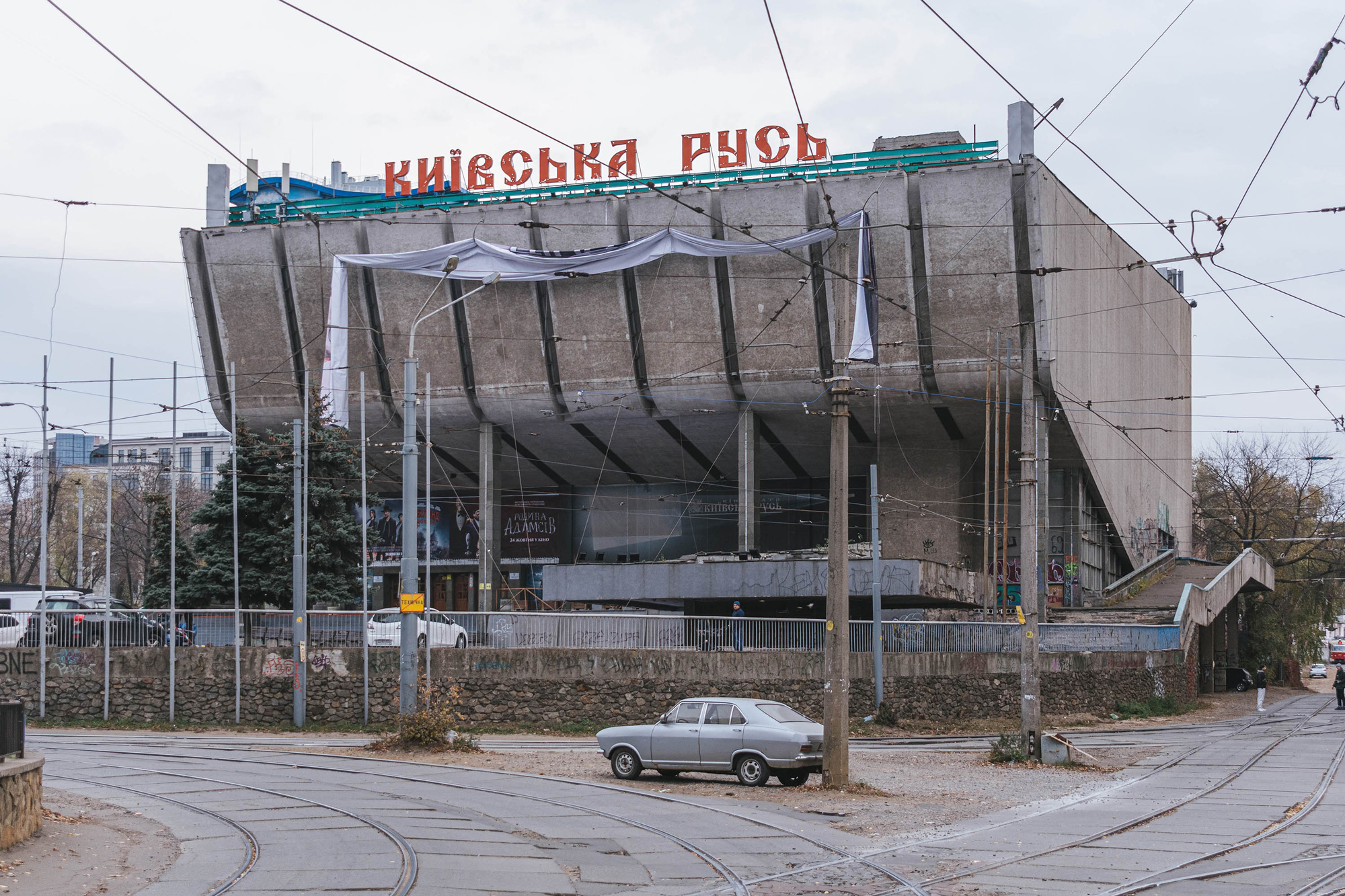 Abandoned concrete building with "київська русь" sign, surrounded by tram tracks and overcast sky.