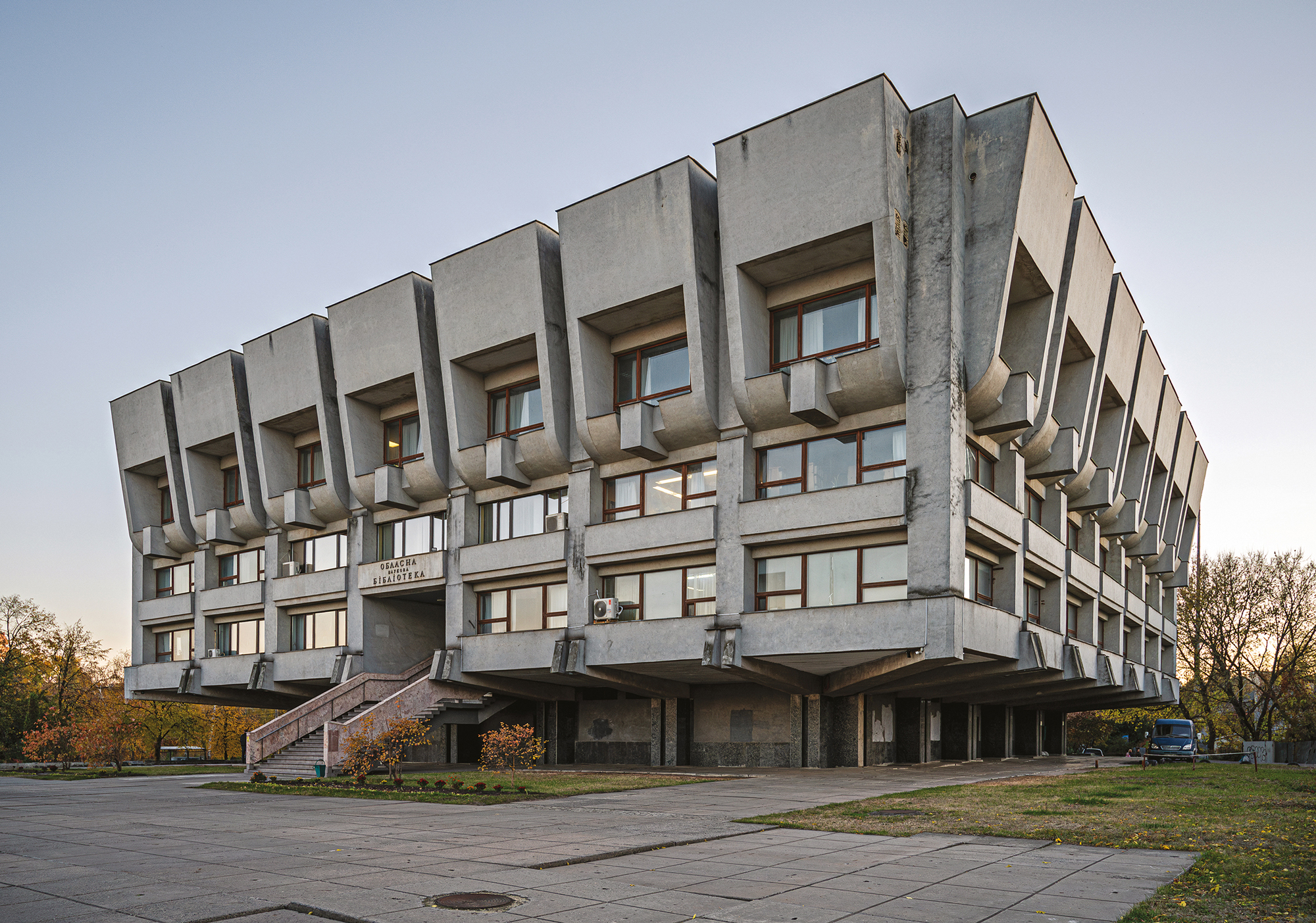 Brutalist-style building with a raised structure and large windows, surrounded by trees and a paved area.