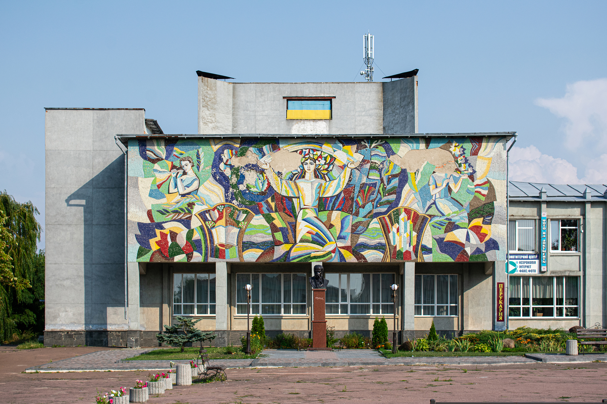 Mural depicting vibrant scenes on the facade of a building, with a statue in front, set in a clear sky.