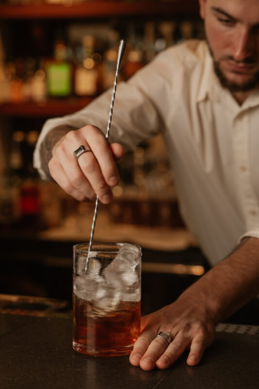 Bartender stirring a cocktail with ice in a glass, surrounded by a blurred bar backdrop.