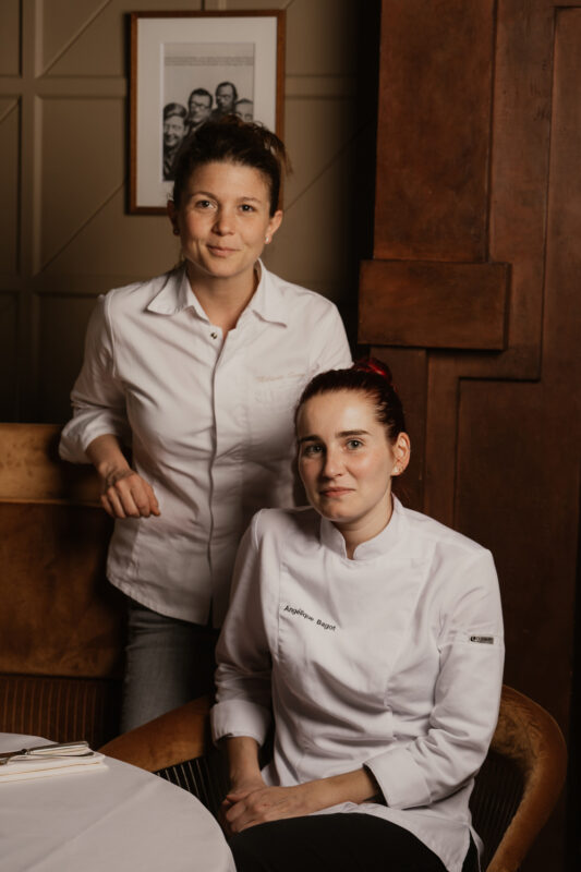 Two women in chef jackets pose together in a restaurant setting, with a framed photo on the wall behind them.