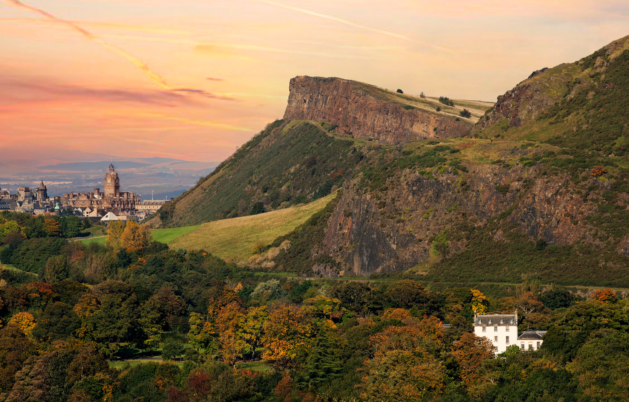 Sunset over Prestonfield House, with hills and colorful foliage in the foreground and a distant city skyline.