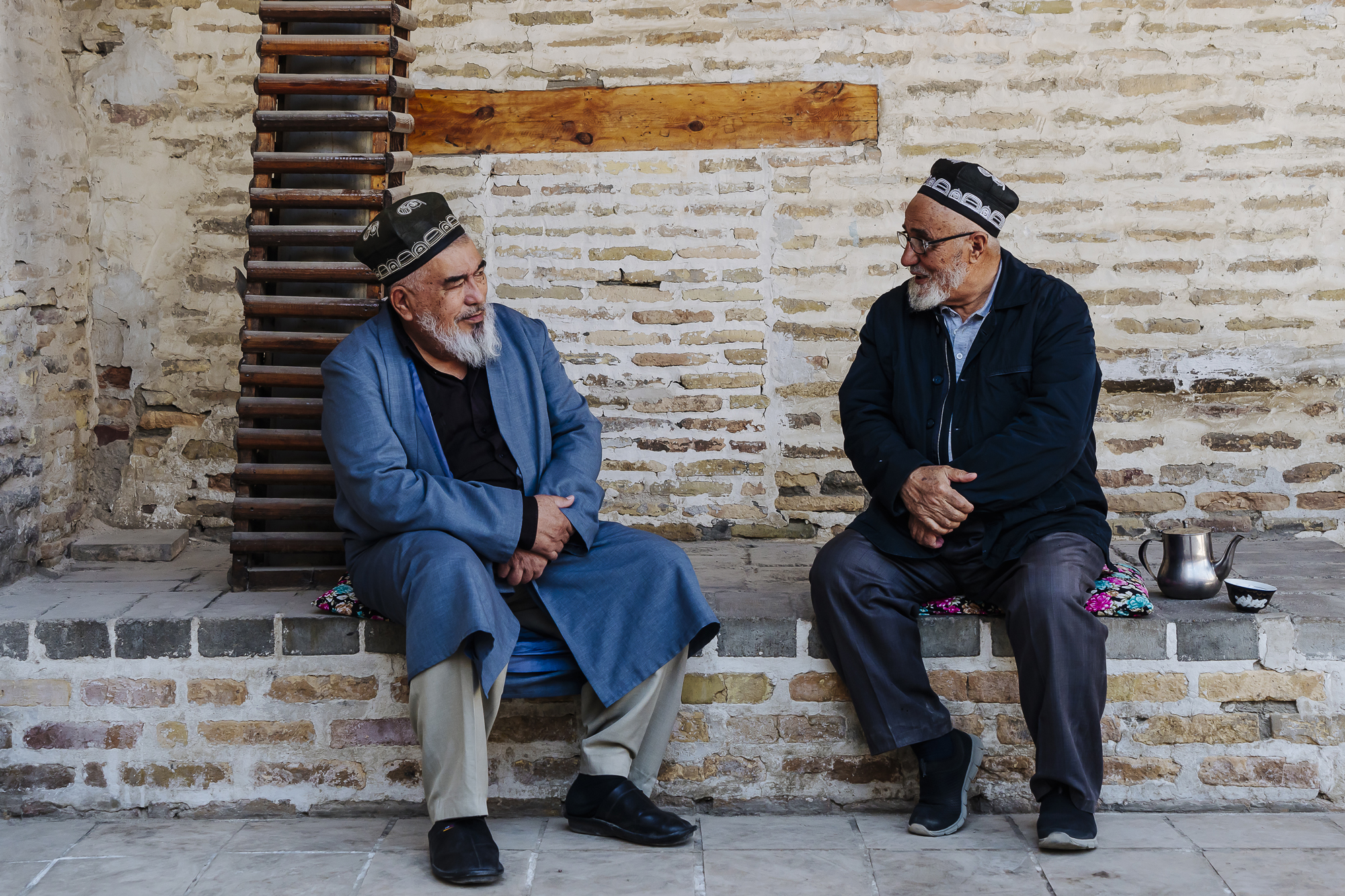 Two elderly men in traditional Uzbek hats sit on a stone bench, engaged in conversation against a brick wall.
