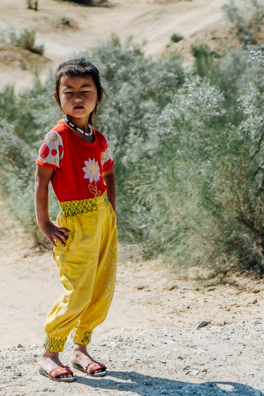 Young girl in a red floral shirt and yellow pants stands confidently in a desert landscape with shrubs.