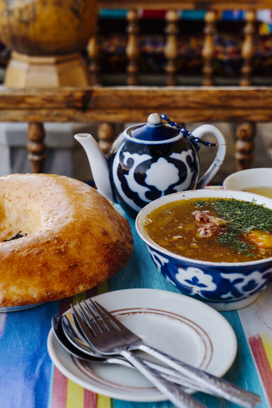 Traditional Uzbek meal featuring a bowl of soup with herbs, a large round bread, and a decorative teapot on a colorful tab...