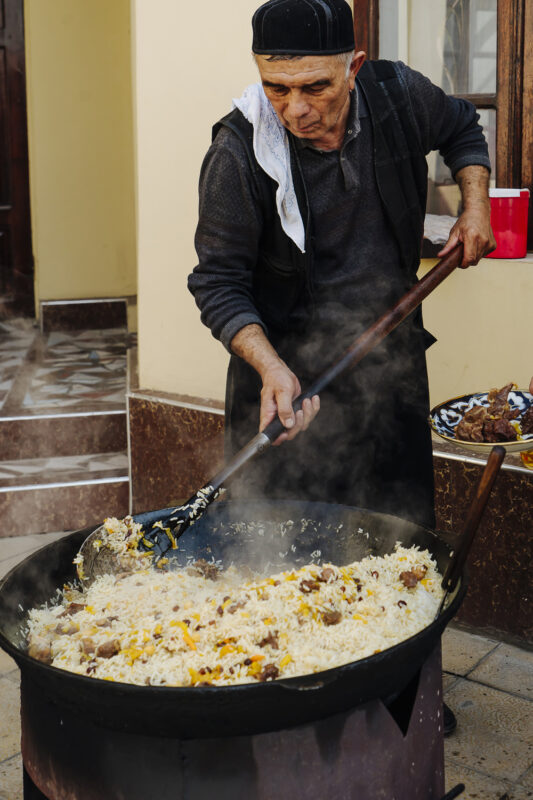 Man cooking pilaf in a large pot outdoors, stirring rice and meat with a wooden spoon, steam rising.