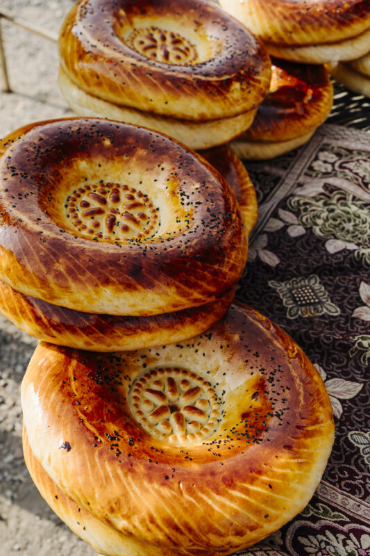 Freshly baked Uzbek bread, stacked with a golden-brown crust and decorative pattern on top, displayed on a patterned cloth.