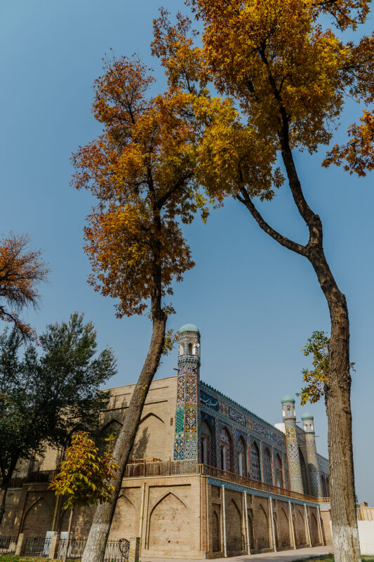 Colorful autumn trees frame a historic building with intricate tilework in Uzbekistan against a clear blue sky.
