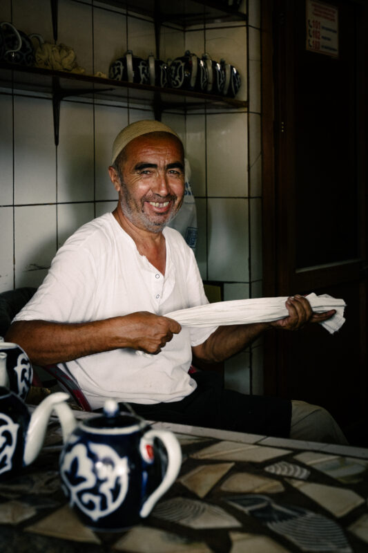 Smiling man in a white shirt holding a cloth, surrounded by traditional ceramic teapots in a dimly lit room.