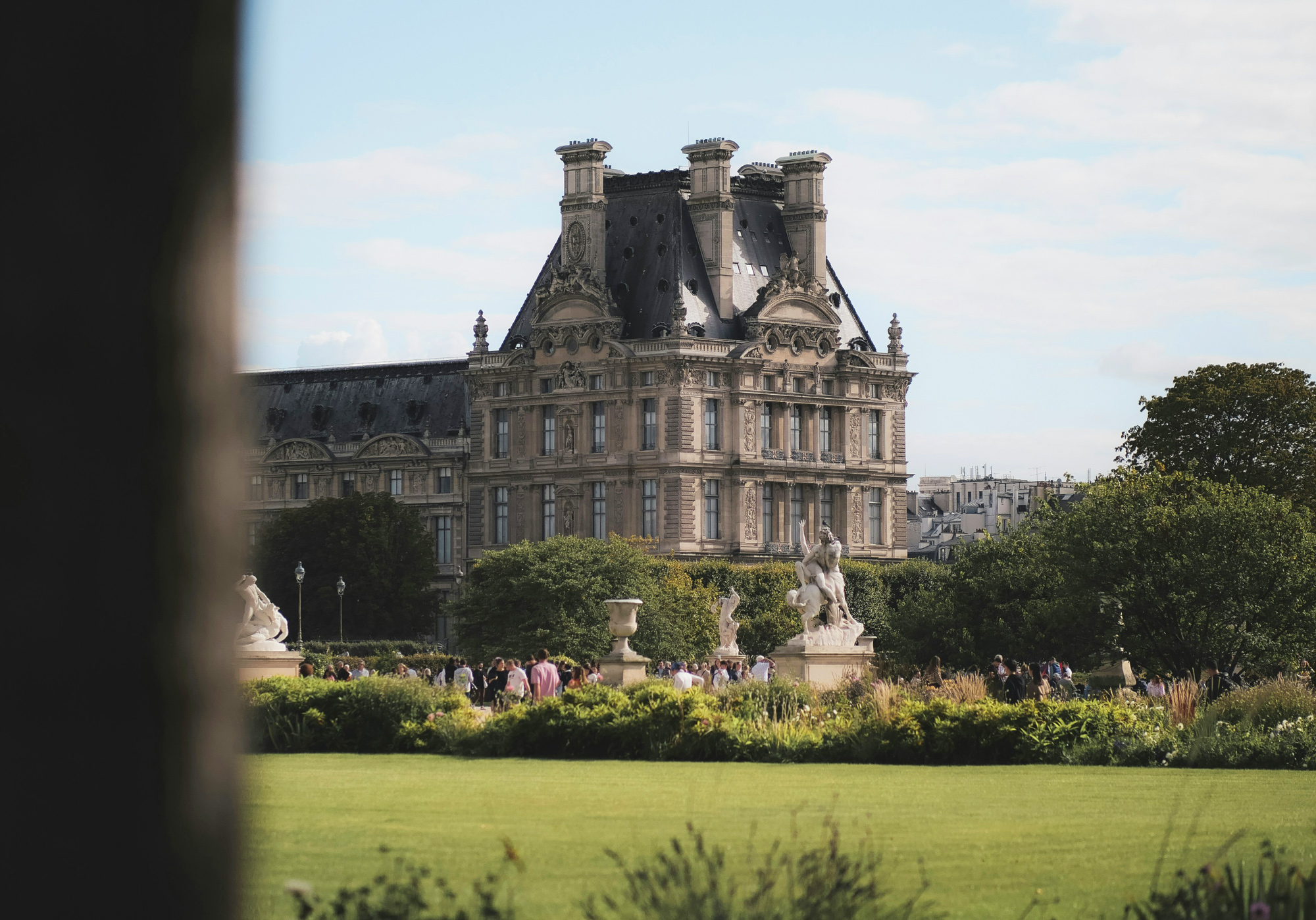 Château Voltaire with sculptures and visitors in the foreground, surrounded by lush greenery and a clear sky.
