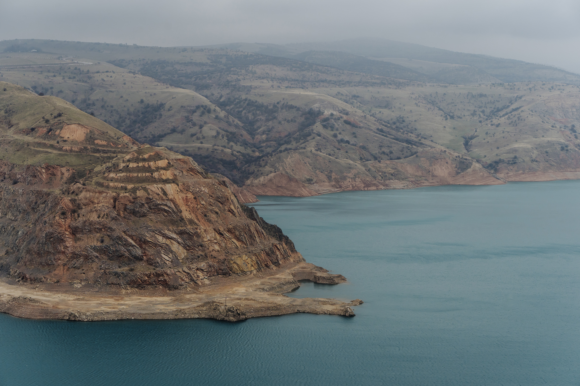 Rocky cliffs along a calm blue lake, surrounded by rolling green hills under a cloudy sky in Uzbekistan.