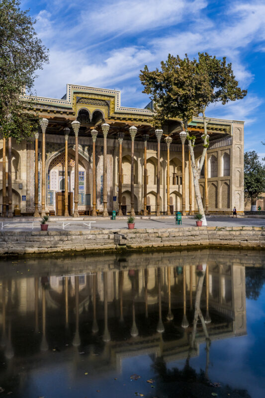 Historic building with ornate columns reflecting in a calm water surface, surrounded by trees and blue sky, titled "бол...