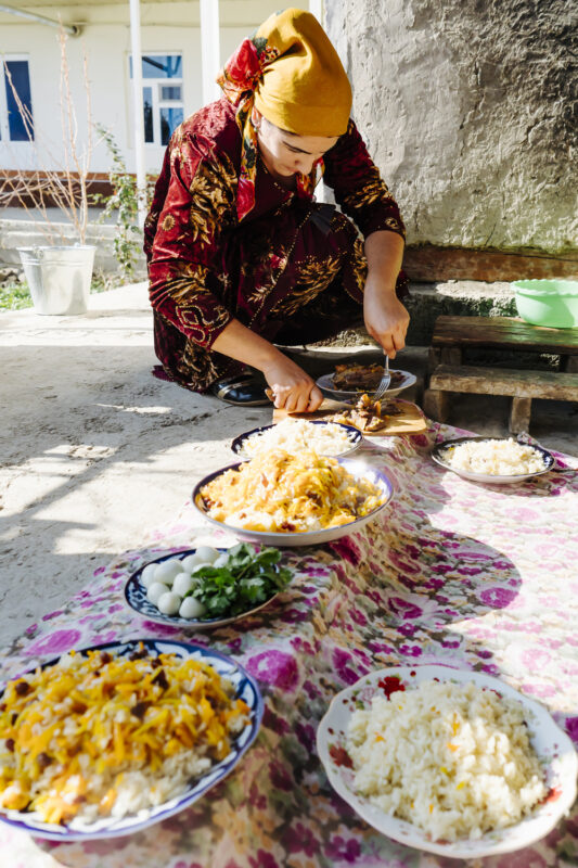 A person in traditional attire prepares plov, surrounded by several plates of the dish and garnishes on a patterned cloth.
