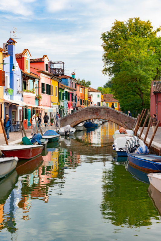 Colorful buildings line a canal in Burano, Venice, with boats moored and a small arch bridge in the background.