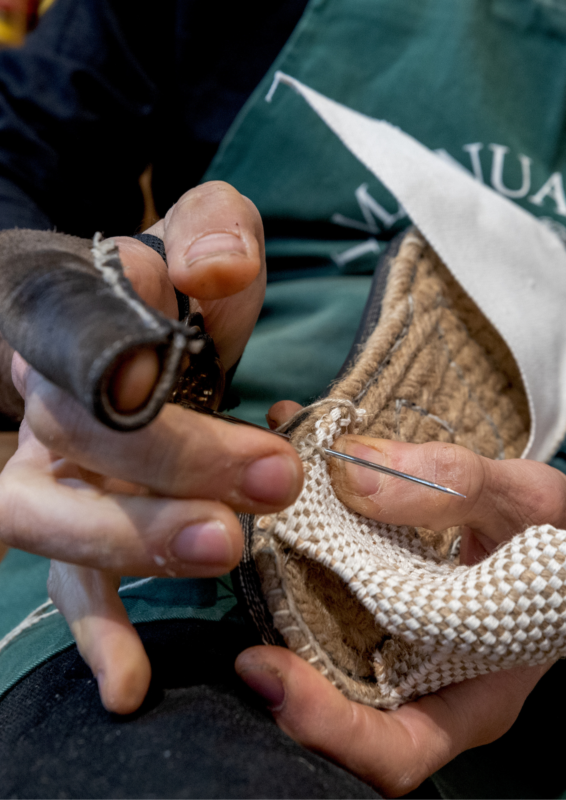 Hands sewing a woven espadrille, showcasing detailed craftsmanship and a needle threading through the material.