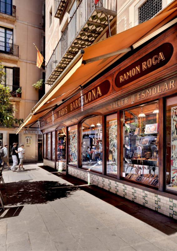 Colorful storefront of "Antigua" in Barcelona, featuring large windows displaying various items, with a sunny street scene.