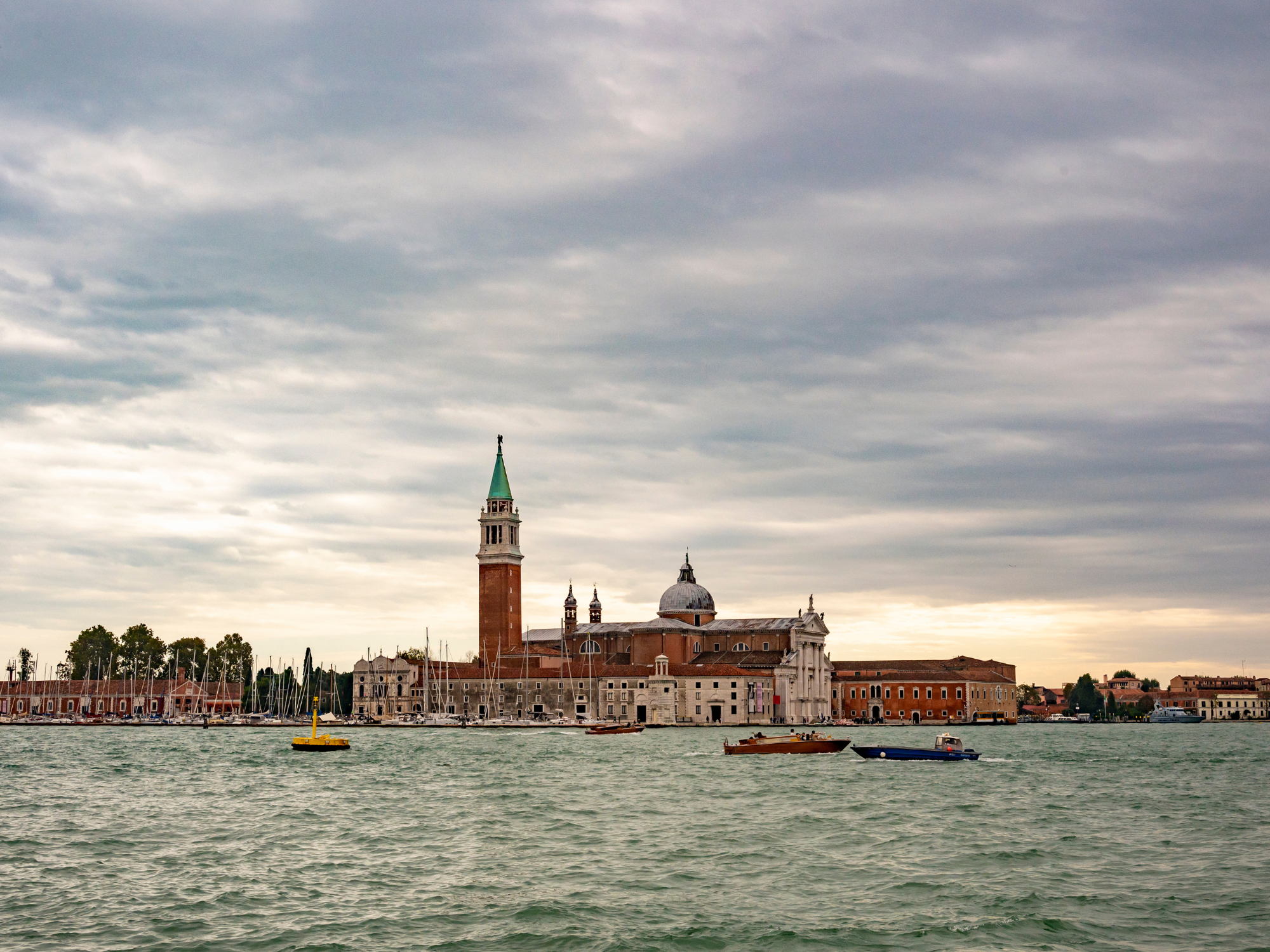 Venetian skyline featuring the bell tower of San Giorgio Maggiore and boats on the water under a cloudy sky.