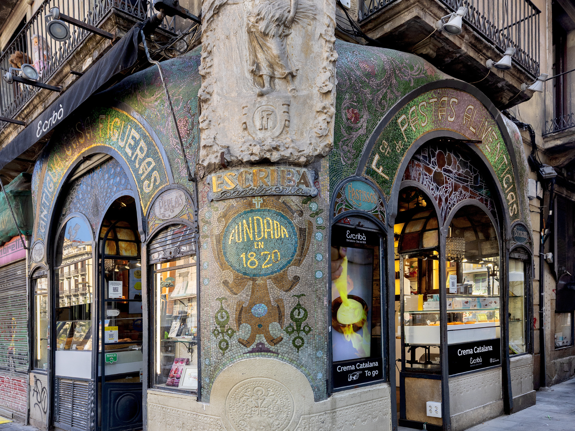 Colorful mosaic-covered corner building in Barcelona, featuring ornate designs and shop windows displaying desserts.