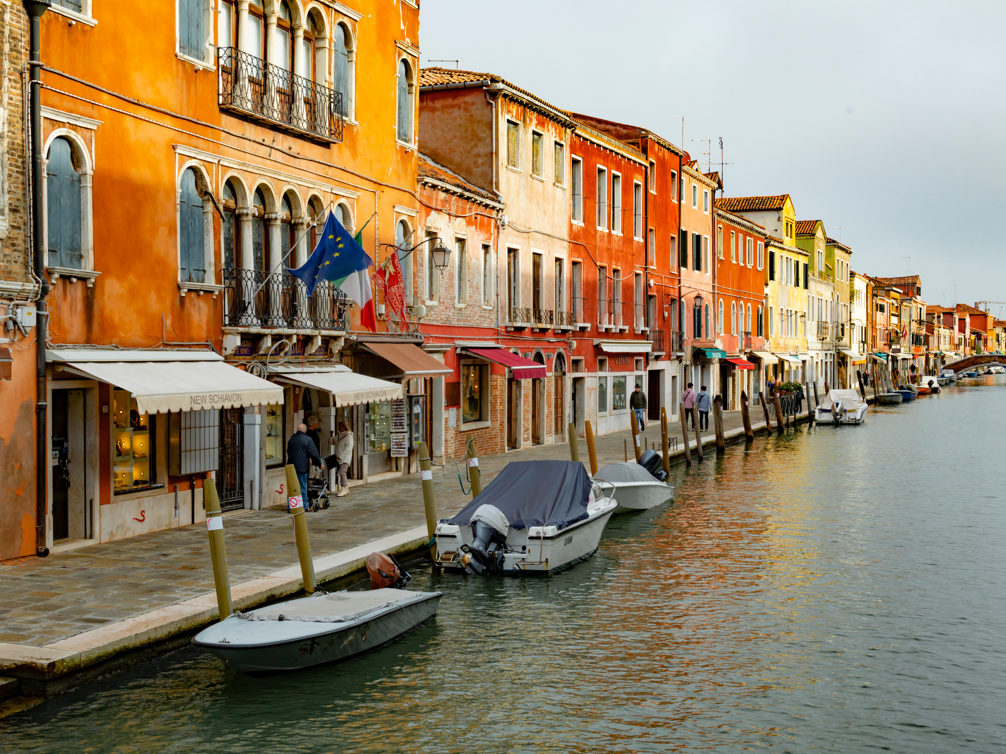 Colorful buildings line a canal in Venice, with boats moored along the waterfront and a flag flying.