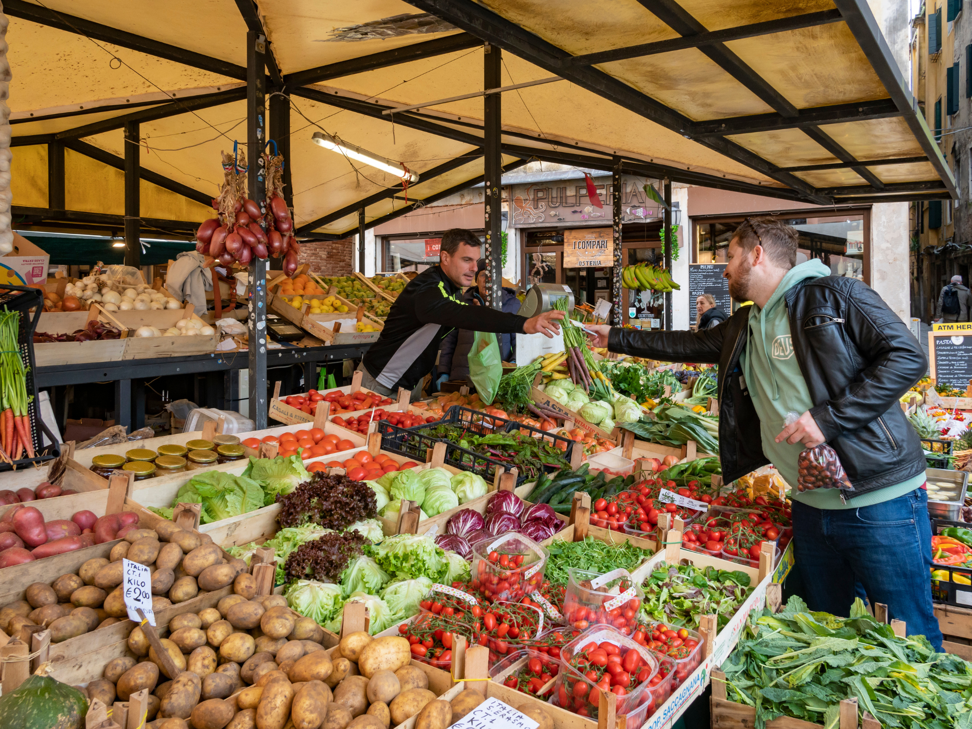 Two men exchanging vegetables at a vibrant market stall filled with fresh produce in Venice.