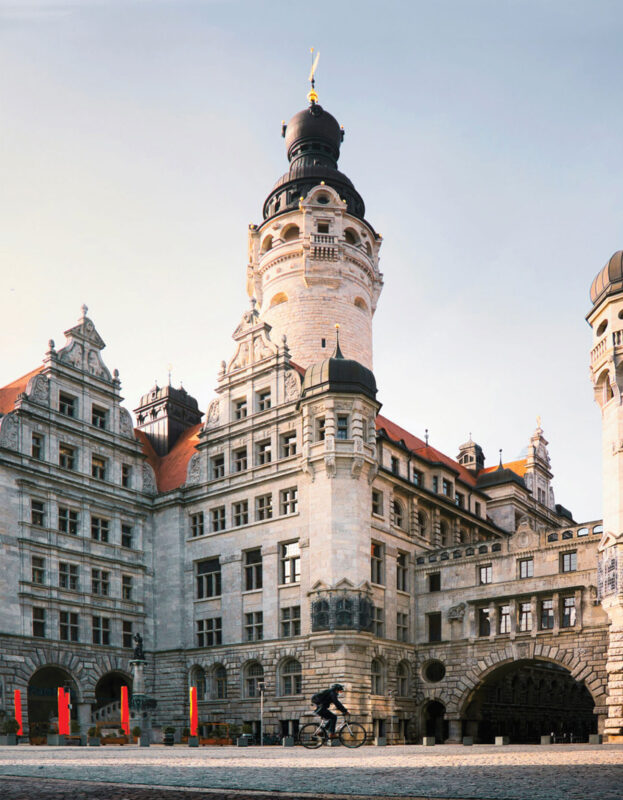 Historic building with a tall tower and ornate architecture, cyclist passing in front, under a clear sky.